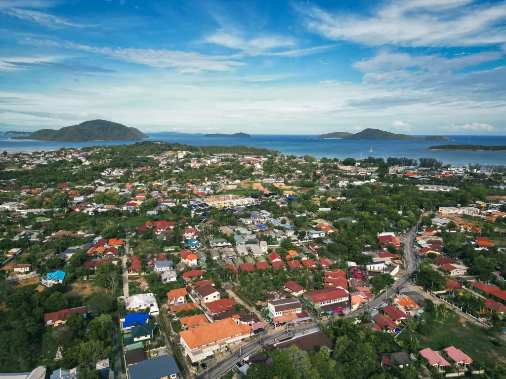 A cinematic aerial view of the sprawling Rawai coastline in Phuket, Thailand. The lush green land meets a deep blue ocean dotted with four distinct islands on the horizon, offering a breathtaking perspective on a future family adventure.